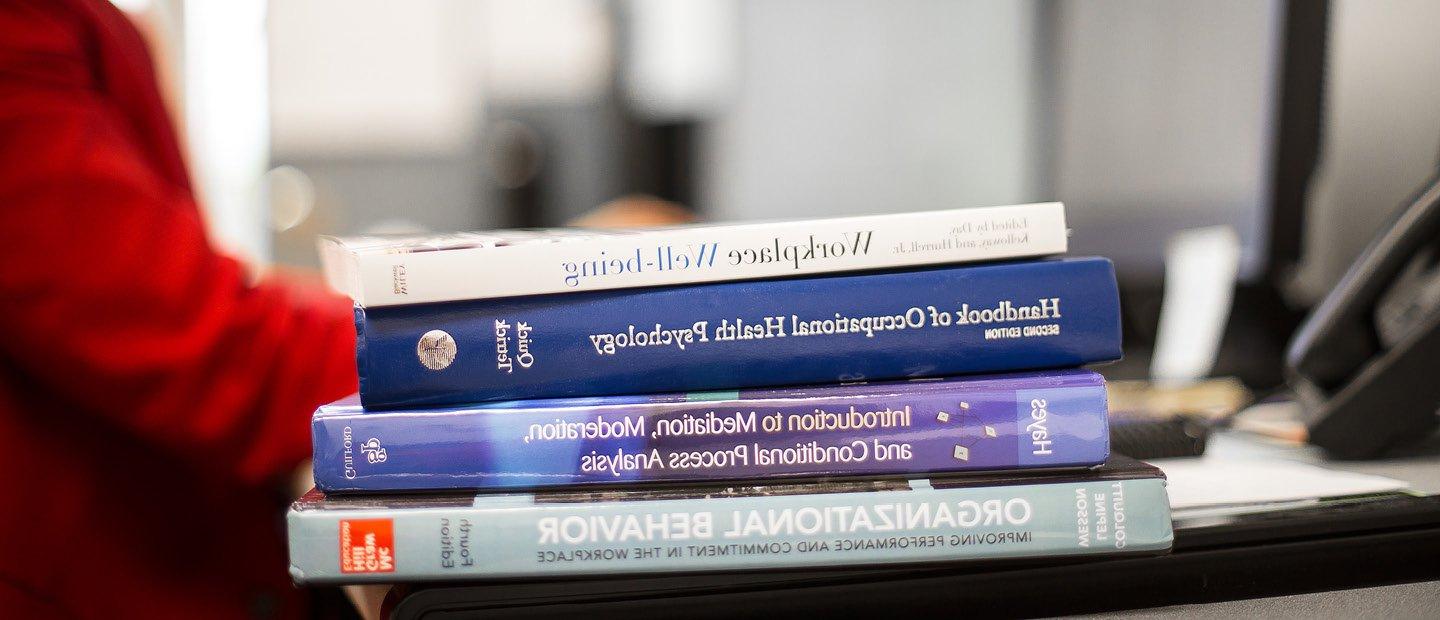 A stack of textbooks on the edge of a desk occupied by a person wearing red.