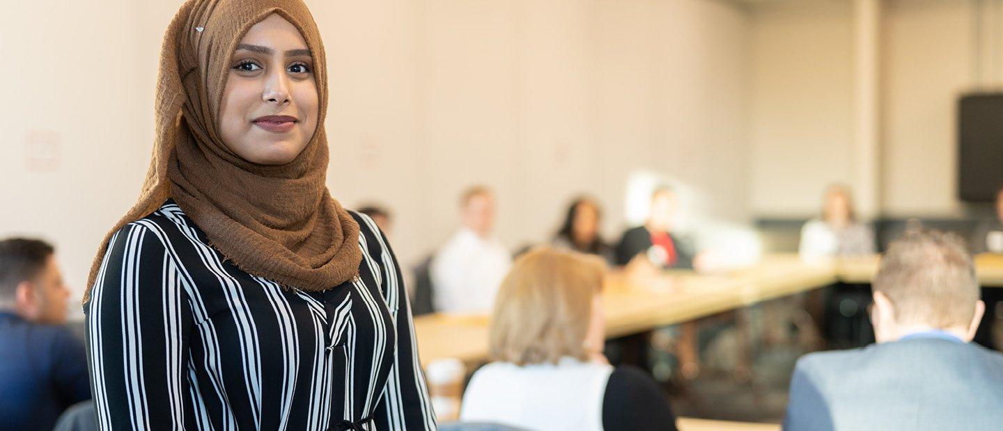 A woman smiling at the camera, standing at the back of a classroom full of people.