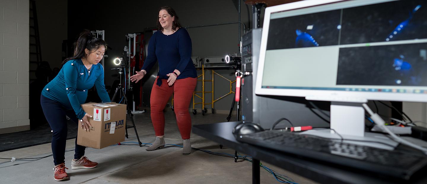 A woman with sensors attached to her body lifts a box, as another woman watches her and demonstrates the motion.